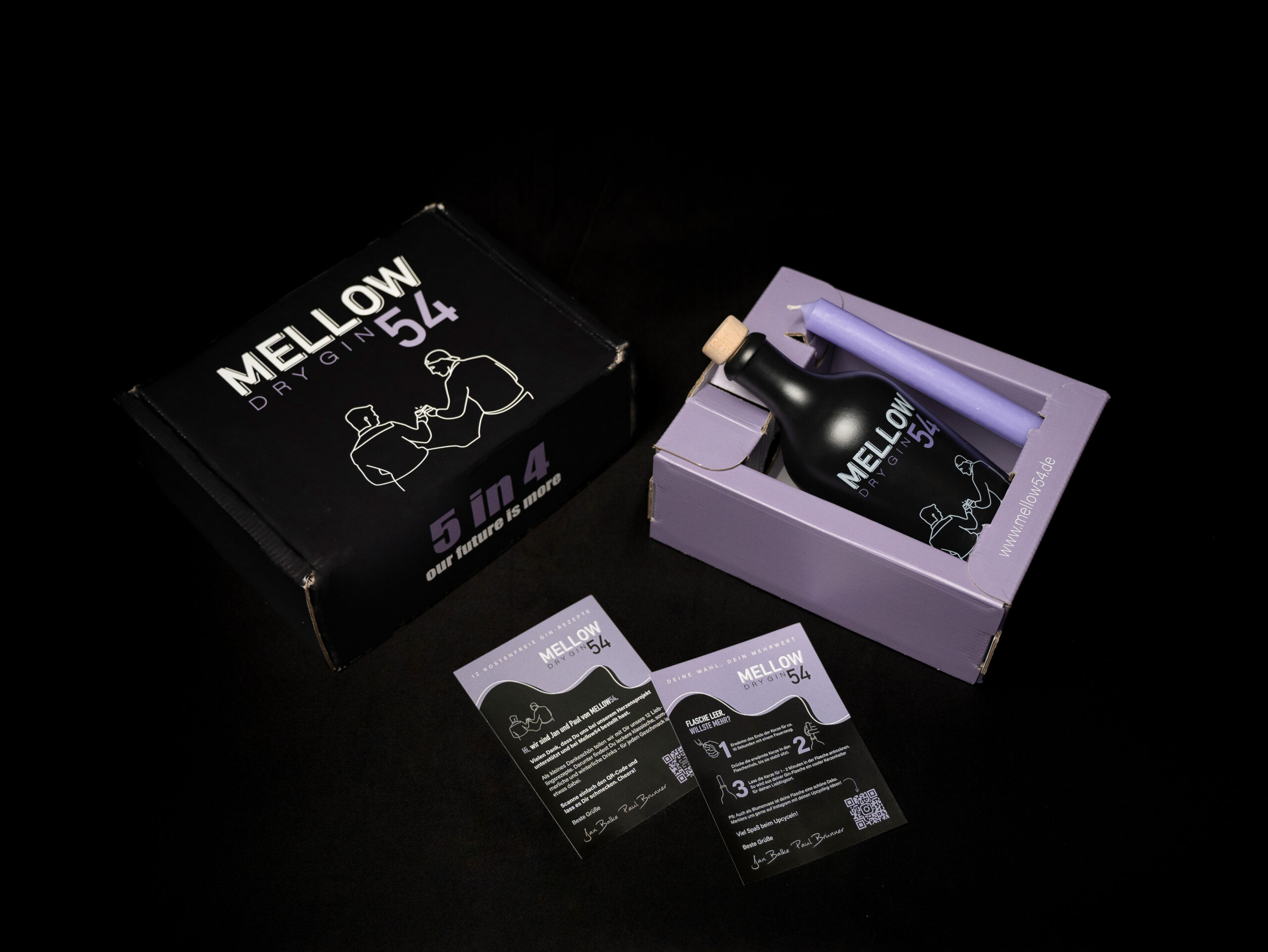 Bild des MELLOW Dry Gin 54 Sets. Das Set enthält eine elegante schwarze Flasche MELLOW Dry Gin 54 mit Korkverschluss, eingebettet in eine stilvolle lavendelfarbene Box. Die Verpackung zeigt minimalistisches Linienkunstwerk von zwei Personen, die Gläser anstoßen, und den Slogan '5 in 4 – our future is more'. Ebenfalls sichtbar sind zwei Informationskarten und eine lavendelfarbene Kerze, alles auf einem dunklen Hintergrund präsentiert. Die Website 'www.mellow54.de' ist auf der Seite der Box aufgedruckt